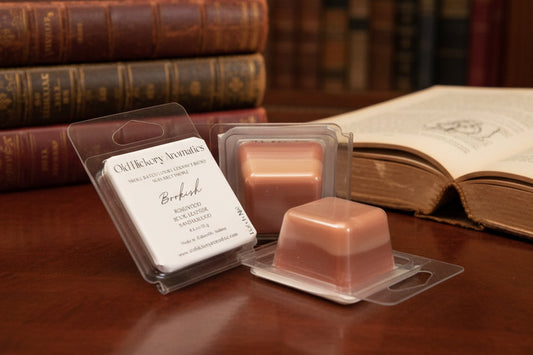 Three sample sized wax melts packaged in clear single cavity clamshell on rosewood table with a stack of leather bound books and one open book in background.