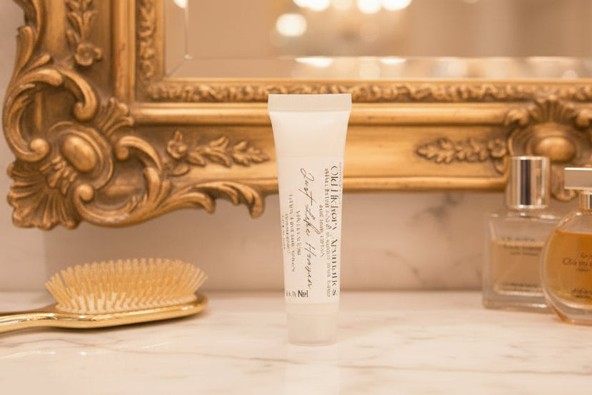Small tube of Old Hickory Aromatics Just Like Heaven Hand and Body Lotion on a marble top table with a gold handled hairbrush, gold framed mirror, and perfume bottles in the background.