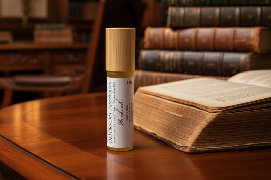 Small rollerball bottle with wooden cap and white label of Old Hickory Aromatics Bookish Roll On Fragrance on a rosewood desk surrounded by leather bound books and a personal library.